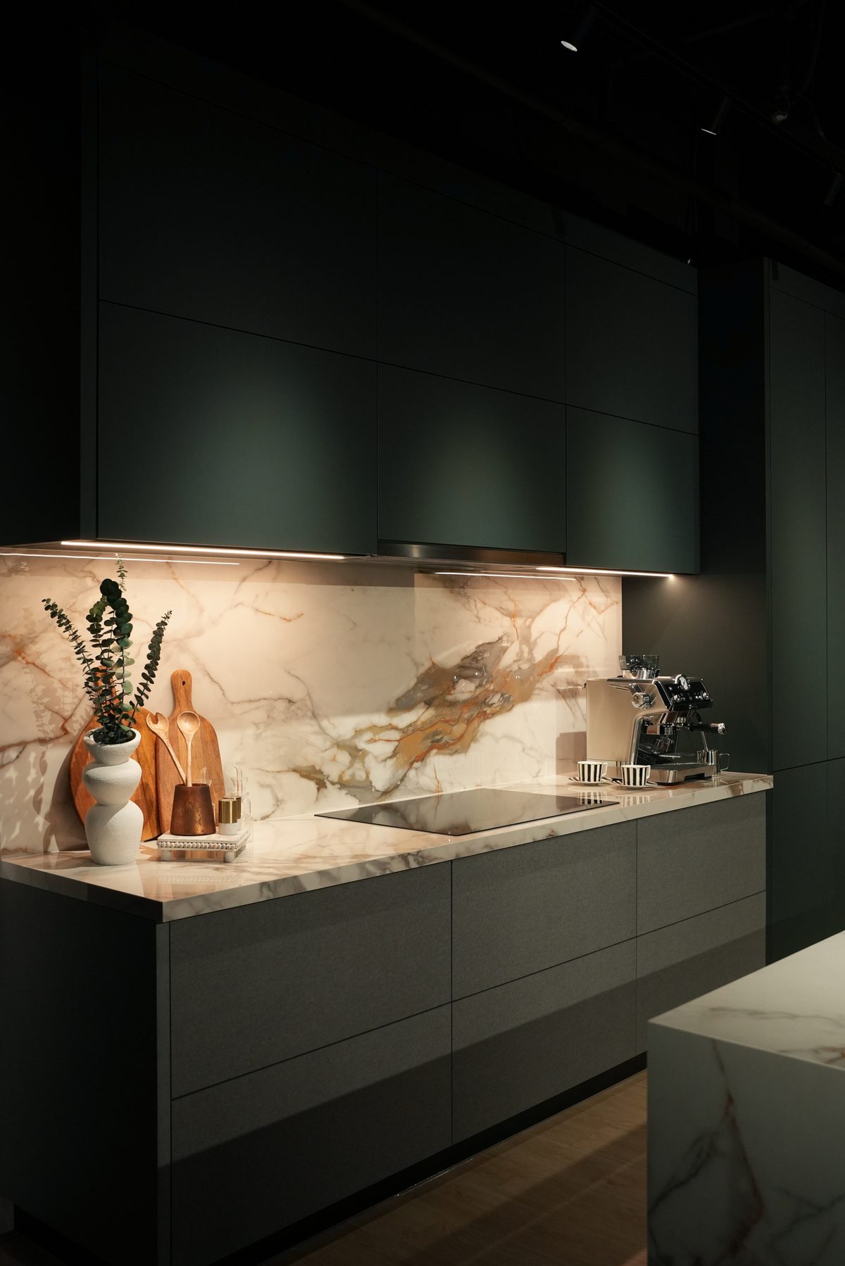 Dark green kitchen with a marble counter and splashback, concealed lighting, and a built-in hob